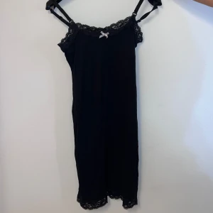 Svart pyjamasklänning med spetsdetaljer - Elegant svart pyjamasklänning med spetsdetaljer vid halsringningen och nederkanten. Klänningen har smala axelband och en söt liten rosett framtill. Perfekt för en stilren look.