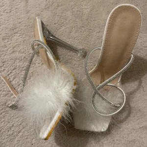 Vita klackskor med fjäderdetalj - Eleganta vita klackskor med transparent klack och glittrande remmar. Skorna har en unik fjäderdetalj framtill som ger en lyxig känsla. Perfekta för speciella tillfällen. Hälhöjd 11.5. Kan gå ner i pris! 