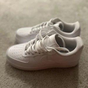 Vita Nike Air Force sneakers - Säljer ett par klassiska vita Nike Air Force sneakers. De har en stilren design med snörning och perforerade detaljer på ovansidan för ventilation. Perfekta för en casual look.