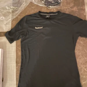 Svart t-shirt från Hummel - Säljer en svart t-shirt från Hummel med korta ärmar och rund hals. Perfekt för träning eller vardagsbruk. T-shirten har en diskret logga på bröstet och är tillverkad i ett bekvämt material.