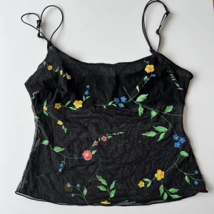 Svart blommig linnetopp - Säljer en svart linnetopp med färgglada blommor i gult, blått och rosa. Toppen har tunna axelband och ett lätt transparent material som ger en luftig känsla. Perfekt för varma dagar!