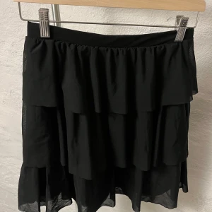 Svart volangkjol - Snygg svart kjol med flera lager av volanger som ger en fin rörelse. Perfekt för att skapa en chic look. Kjolen har en elastisk midja för bekväm passform.