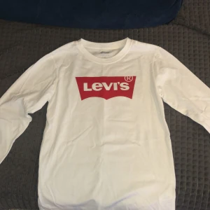 Vit långärmad t-shirt från Levi's - Säljer en klassisk vit långärmad t-shirt från Levi's med det ikoniska röda logotyptrycket på bröstet. Perfekt för en avslappnad stil.