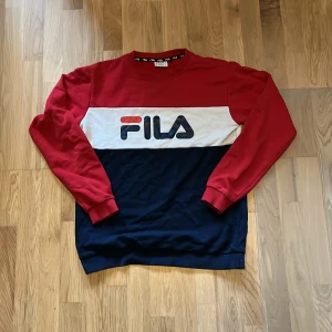 Flerfärgad sweatshirt från FILA - Säljer en snygg flerfärgad sweatshirt från FILA med rött, vitt och blått blockmönster. Tröjan har långa ärmar och ett stort FILA-tryck på framsidan. Perfekt för en sportig look!