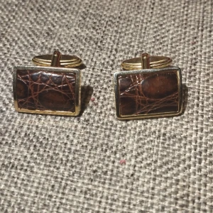 Bruna manschettknappar med gulddetaljer - Snygga rektangulära manschettknappar krokodil imitation med en brun yta och guldfärgad ram. Perfekta för att ge en elegant touch till din outfit. Nytt skick dock inte finns någon förpackning eller etikett.