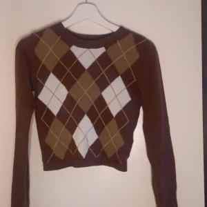 Brun tröja med argylemönster från Hollister - Säljer en brun tröja från Hollister med klassiskt argylemönster i brunt och vitt. Tröjan har långa ärmar och en croppad passform, perfekt för en stilren look. Tillverkad i mjukt material för extra komfort.