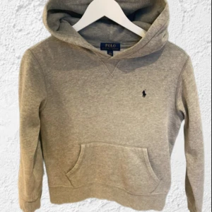 Grå hoodie från Polo Ralph Lauren - Säljer en klassisk grå hoodie från Polo Ralph Lauren med en liten broderad logga på bröstet. Tröjan har en praktisk magficka och en bekväm huva. Perfekt för en avslappnad stil.