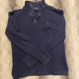 Blå tröja från Ralph Lauren - Säljer en stilren klassisk marinblå quarter zip av Ralphlauren. Perfekt nu inför mildare tider. Storlek 14-16 i barn storlek. Modell är 170cm. Tröjan har en liten genom slinkig fläck på höger armled men är över huvud taget knappast synbar.