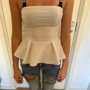 Vit korsettliknande topp - Säljer en snygg vit korsettliknande topp med peplumdetalj. Toppen har smala axelband och en tight passform som framhäver figuren. Perfekt för festliga tillfällen!