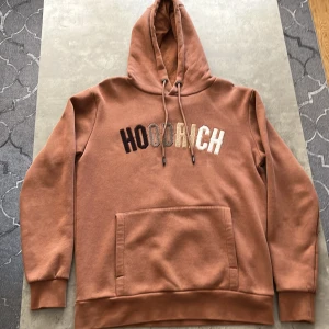 Brun hoodie från Hoodrich - Säljer en snygg brun hoodie från Hoodrich i storlek S. Den har en stor ficka fram och texten 'HOODRICH' broderad i olika färger på bröstet. Perfekt för höst och vinter med sin mysiga och varma känsla. Passar både till vardags och chillkvällar.