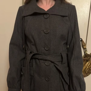 Mörkgrå kappa från Vila - Säljer en snygg mörkgrå kappa från Vila i storlek L. Den har långa ärmar och knäpps med stora knappar framtill. Kappan har ett bälte i midjan för en fin passform och är perfekt för höst och vinter. Den är i nyskick och redo för nya äventyr!