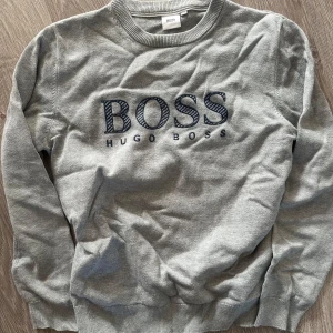 Grå tröja från Hugo Boss - Säljer en stilren grå tröja från Hugo Boss. Den har en klassisk design med BOSS-loggan broderad på framsidan. Tröjan är långärmad och perfekt för både vardag och lite finare tillfällen. Superbekväm och i bra skick!