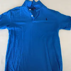 Säljer en snygg blå pikétröja från Polo Sport Ralph Lauren. Den har en klassisk krage med två knappar och det ikoniska logotypbroderiet på bröstet. Perfekt för en avslappnad men stilren look!