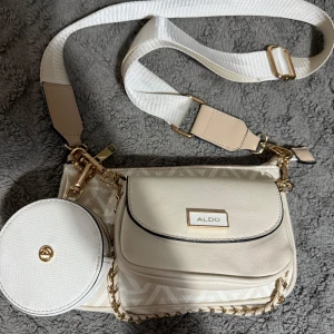 Beige axelväska från Aldo - Säljer en superfin beige axelväska från Aldo. Den har ett snyggt mönster och en justerbar axelrem. Väskan har både dragkedja och en liten rund väska framtill som extra detalj. Perfekt för både vardag och fest!