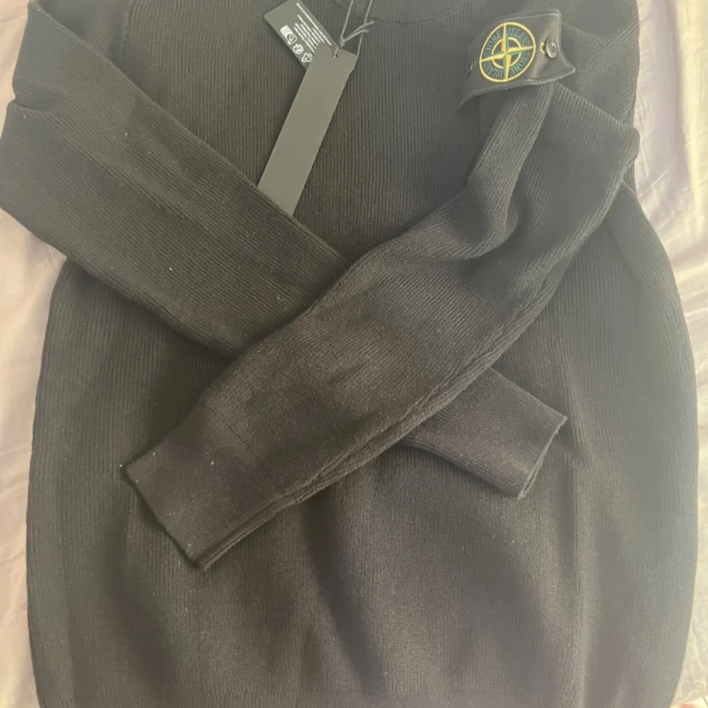 Säljer en snygg svart stickad tröja från Stone Island. Den har en klassisk design med långärmade ärmar och en ribbad struktur. Perfekt för höst och vinter, och den ikoniska loggan på ärmen ger en extra touch av stil.. Tröjor & Koftor.