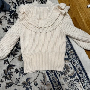 Beige stickad tröja med volangdetaljer - Säljer en snygg beige stickad tröja med volangdetaljer på axlarna. Tröjan har långa ärmar och en ribbad design som ger en mysig känsla. Perfekt för kyligare dagar!