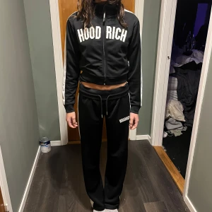 Svart träningsset från Hoodrich XS *NYTT* - Helt nytt snyggt svart träningsset från Hoodrich med vita detaljer. Jackan har dragkedja och texten 'Hoodrich' tryckt på bröstet. Byxorna har en matchande vit rand längs sidan och samma tryck på benet. Perfekt för en sportig look! Endast provat!!