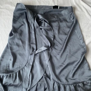 Silvrig kjol från Neo Noir - Snygg omlottkjol från Neo Noir med volangdetaljer. Kjolen har en knytning framtill. Aldrig använd. 