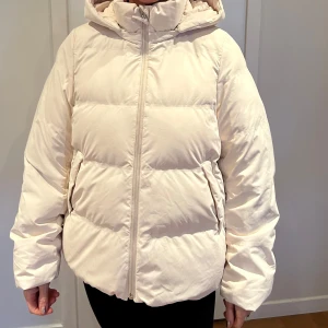 Beige pufferjacka från Uniqlo - Säljer en beige pufferjacka från Uniqlo. Jackan har en dragkedja framtill och en mysig huva. Perfekt för kyliga dagar med sin vadderade design.