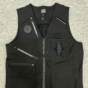 Svart väst från Black Squad - Snygg svart väst från Black Squad med dragkedja och flera praktiska fickor. Västen har en cool design med textdetaljer och spänne framtill. Perfekt för en urban look.