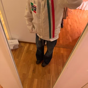 Vit jacka med Castrol-tryck thrifted  - Säljer en cool vit jacka med Castrol GTX-tryck och färgglada ränder i rött och grönt. Jackan har en broderad detalj på ärmen med texten 'Rally Marshals Association'. Perfekt för bilentusiaster!