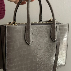 Grå krokodilpräglad handväska - Snygg grå handväska med krokodilpräglat mönster och guldfärgade dragkedjor har aldrig används från hm gammal så finns ej i butik mer jätte söt och fin men har kommit fram till att små väskor är min grej💗