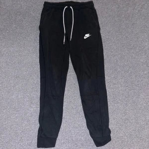 Svarta mjukisbyxor från Nike - Säljer ett par svarta mjukisbyxor från Nike med vit snörning i midjan och logga på benet. Byxorna har en bekväm passform och är perfekta för avslappnade dagar.