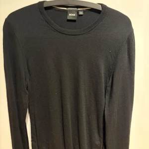 Svart tröja från BOSS - Säljer en stilren svart tröja från BOSS, tillverkad av finaste italienska garn. Tröjan har en klassisk rund hals och långa ärmar, perfekt för en elegant look. Passar utmärkt till både vardag och mer formella tillfällen.