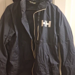 Svart vindjacka från Helly Hansen - Snygg svart vindjacka från Helly Hansen med dragkedja och huva. Perfekt för blåsiga dagar. Jackan har en enkel design med HH-logga på bröstet.