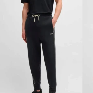 Svarta mjukisbyxor från HUGO BOSS - Snygga svarta mjukisbyxor från BOSS med vit snörning i midjan och diskret logga på sidan. Byxorna har en avslappnad passform och vita ränder längs sidorna för en sportig look.