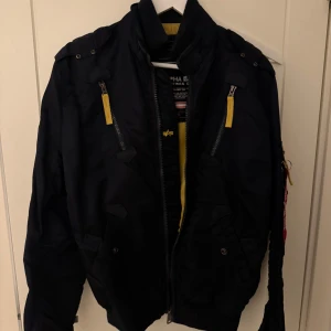 Mörkblå bomberjacka från Alpha Industries - Säljer en mörkblå bomberjacka från Alpha Industries med gulddetaljer och broderad logga på ärmen. Jackan har dragkedja och flera fickor, inklusive en med 'Remove Before Flight'-tag. Perfekt för en cool och avslappnad stil. Använd ett fåtal gånger 