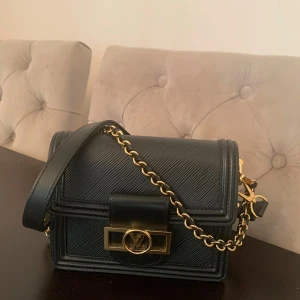 Svart axelväska från Louis Vuitton - Elegant svart axelväska från Louis Vuitton med guldfärgade detaljer och en kedja. Väskan har en stilren design med ett klassiskt LV-spänne framtill. Perfekt för att ge en lyxig touch till din outfit.