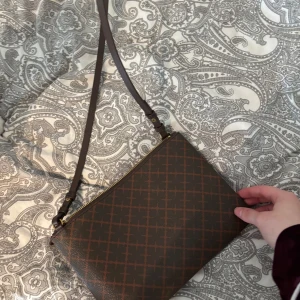 By Malene birger - Snygg brun axelväska med ett diskret rutigt mönster och guldfärgad dragkedja. Väskan har en justerbar axelrem och är perfekt för att bära dina nödvändigheter. Insidan är ljus och rymlig.