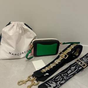 Grön och svart axelväska från Marc Jacobs - Snygg axelväska från Marc Jacobs i en cool kombination av grönt och svart. Väskan har en justerbar axelrem med guldiga detaljer och en praktisk dragkedja. Perfekt för att ge din outfit en färgklick!