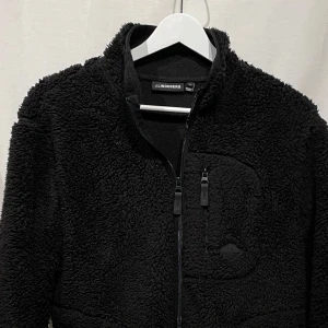 Svart fleecejacka från J.lindeberg - Mysig svart fleecejacka med dragkedja. Perfekt för kyliga dagar och har en praktisk bröstficka. Jackan är långärmad och har en enkel, stilren design. Modellen är 180cm