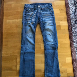 True religion jeans - Säljer ett par blå jeans med slitningar och en trasig detalj på framsidan. De har en klassisk femficksdesign och knappar bak. Perfekta för en avslappnad stil. True religion byxor.