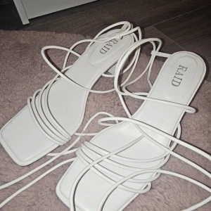 Vita sandaler från RAID - Stilrena vita sandaler från RAID med smala remmar och fyrkantig tå. Perfekta för sommarens alla tillfällen. Sandalerna har en elegant design med långa snören som kan knytas runt ankeln.