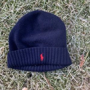 Svart mössa från Polo Ralph Lauren - Snygg mörk blå  mössa från Polo Ralph Lauren med röd broderad logga framtill. Perfekt för kyliga dagar med sin ribbade kant som ger extra värme och stil.storleken är 43cm men kan töjas