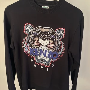 Svart tröja med tigerbroderi från Kenzo - Snygg svart tröja från Kenzo med ett ikoniskt tigerbroderi i flera färger på framsidan. Tröjan har långa ärmar och en rund halsringning. Perfekt för att ge din outfit en cool touch.nypris 3500kr