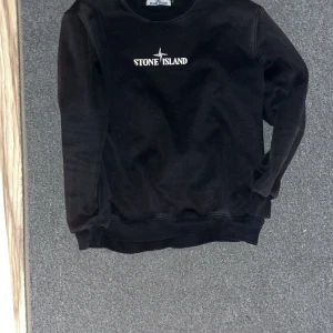  Stone Island Tröja  - Snygg svart sweatshirt från Stone Island med logga på bröstet och stort tryck på ryggen. Tröjan har långa ärmar och ribbade muddar vid ärmslut och nederkant. Perfekt för en stilren och avslappnad look.