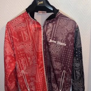 Mönstrad jacka från Palm Angels - Säljer min tröja  från Palm Angels med ett unikt paisleymönster i rött och lila. Jackan har dragkedja och långa ärmar med vita ränder längs ärmarna. Perfekt för att sticka ut i mängden! Storlek M ✅📦
