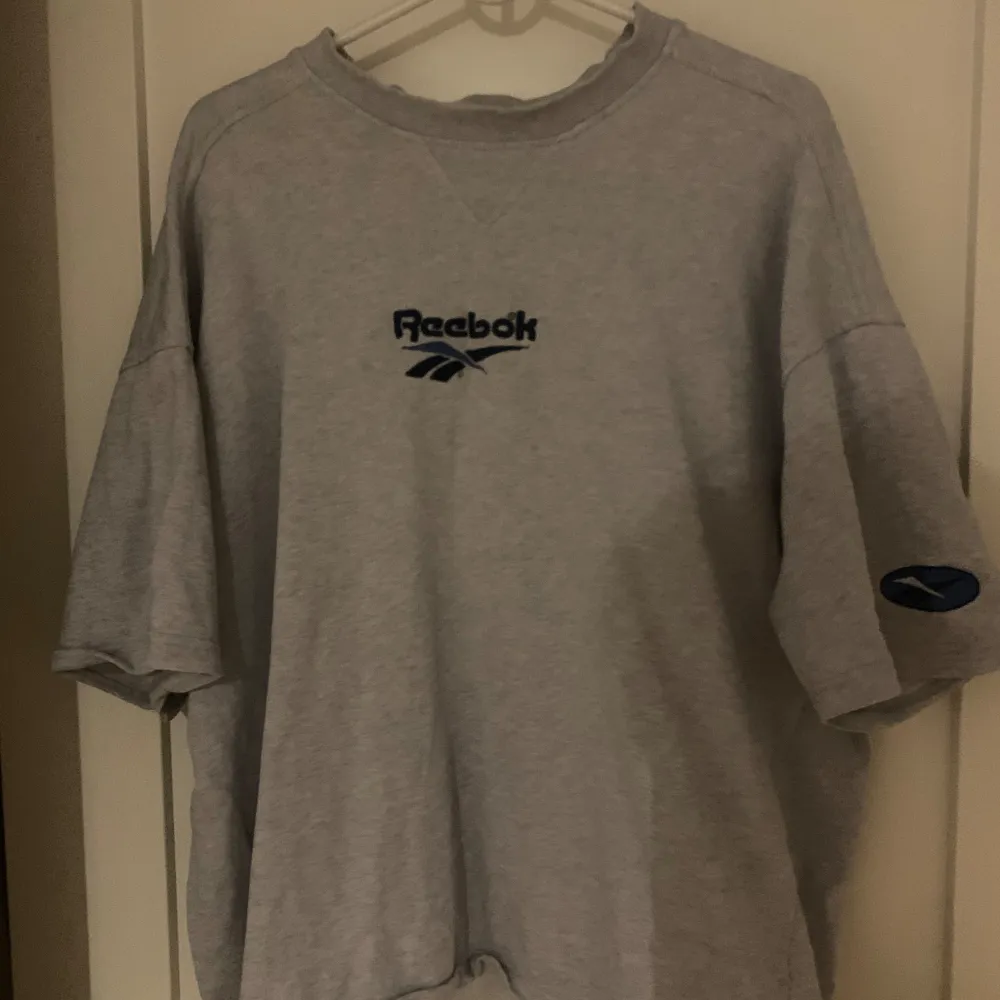 Säljer en grå t-shirt från Reebok med deras klassiska logga i blått på bröstet. T-shirten har korta ärmar och en rund halsringning. Baggy och vintage . T-paidat.