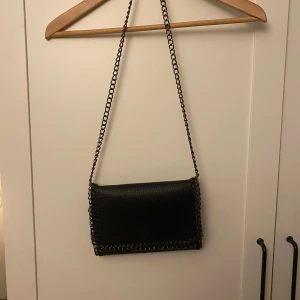Svart axelväska med kedjedetaljer - Snygg svart axelväska i skinn med kedjedetaljer runt kanterna.