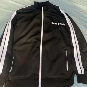 Svart träningsjacka från Palm Angels - Snygg svart träningsjacka från Palm Angels med vita ränder längs ärmarna och dragkedja framtill. Jackan har en hög krage och två fickor med dragkedja. Perfekt för en sportig look.