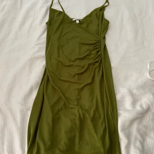 Olivgrön omlottklänning från H&M - Säljer en superfin olivgrön omlottklänning från H&M. Den har smala axelband och en snygg drapering på sidan. 