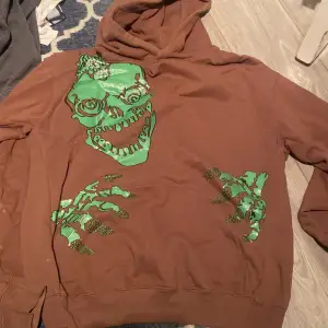 Säljer en cool brun hoodie med ett grönt dödskalletryck på framsidan. Den har långa ärmar och en klassisk huva. Perfekt för höst och vinter när du vill ha något bekvämt men ändå stiligt. Passar bra till både jeans och joggers!