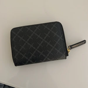 Svart plånbok med dragkedja - Snygg svart plånbok från By Malene Birger med ett diskret mönster och praktisk dragkedja. Insidan är i en ljusare nyans med flera fack för kort och kontanter. Perfekt för att hålla ordning på dina värdesaker.