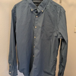 Blå skjorta från Tommy Hilfiger - Snygg blå skjorta från Tommy Hilfiger med diskret mönster och klassisk krage. Skjortan har långa ärmar och knappar framtill samt en bröstficka med logotypdetalj. Perfekt för en stilren look.