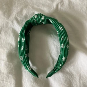 Grönt diadem med blommönster - Snyggt grönt diadem med vita blommor. Perfekt för att ge en färgklick till din outfit. Diademet har en knutdetalj på toppen som ger en extra touch.