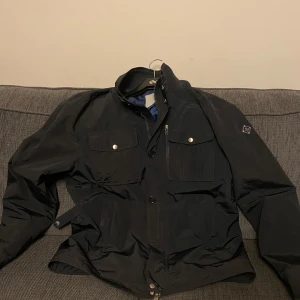 Svart jacka med knappar - Säljer en svart jacka med knappar och flera fickor. Jackan har en stilren design med en hög krage och är perfekt för kyligare dagar. Den har en klassisk look som funkar till det mesta.